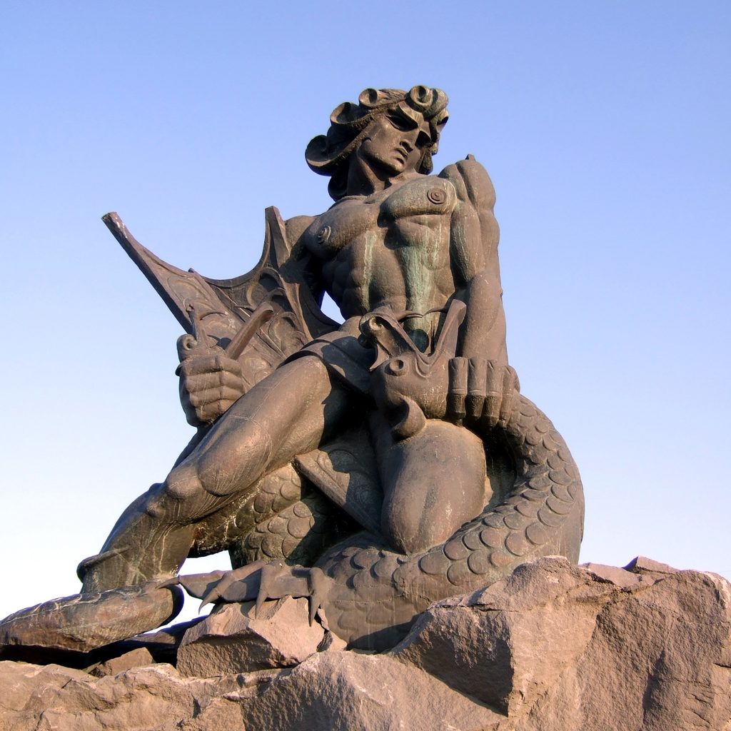 A14_Image | THE ANCIENT WORLD Stone statue of a winged mythical creature perched on a rocky pedestal against a clear blue sky.